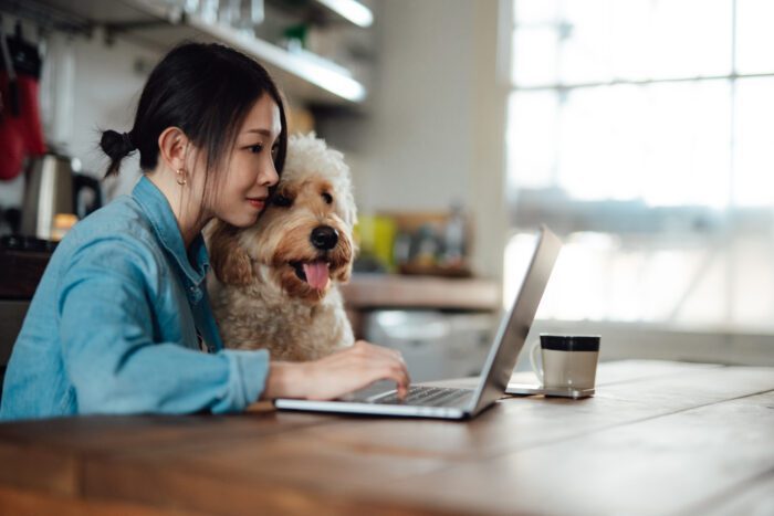 Young Asian woman using laptop next to her dog, sitting at dining table at home. Work life balance. Living with a pet. Online shopping at home.