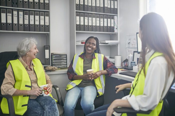 Female workers laughing while sitting in carpet factory. Happy colleagues are communicating during coffee break. Coworkers are wearing reflective clothing in industry.