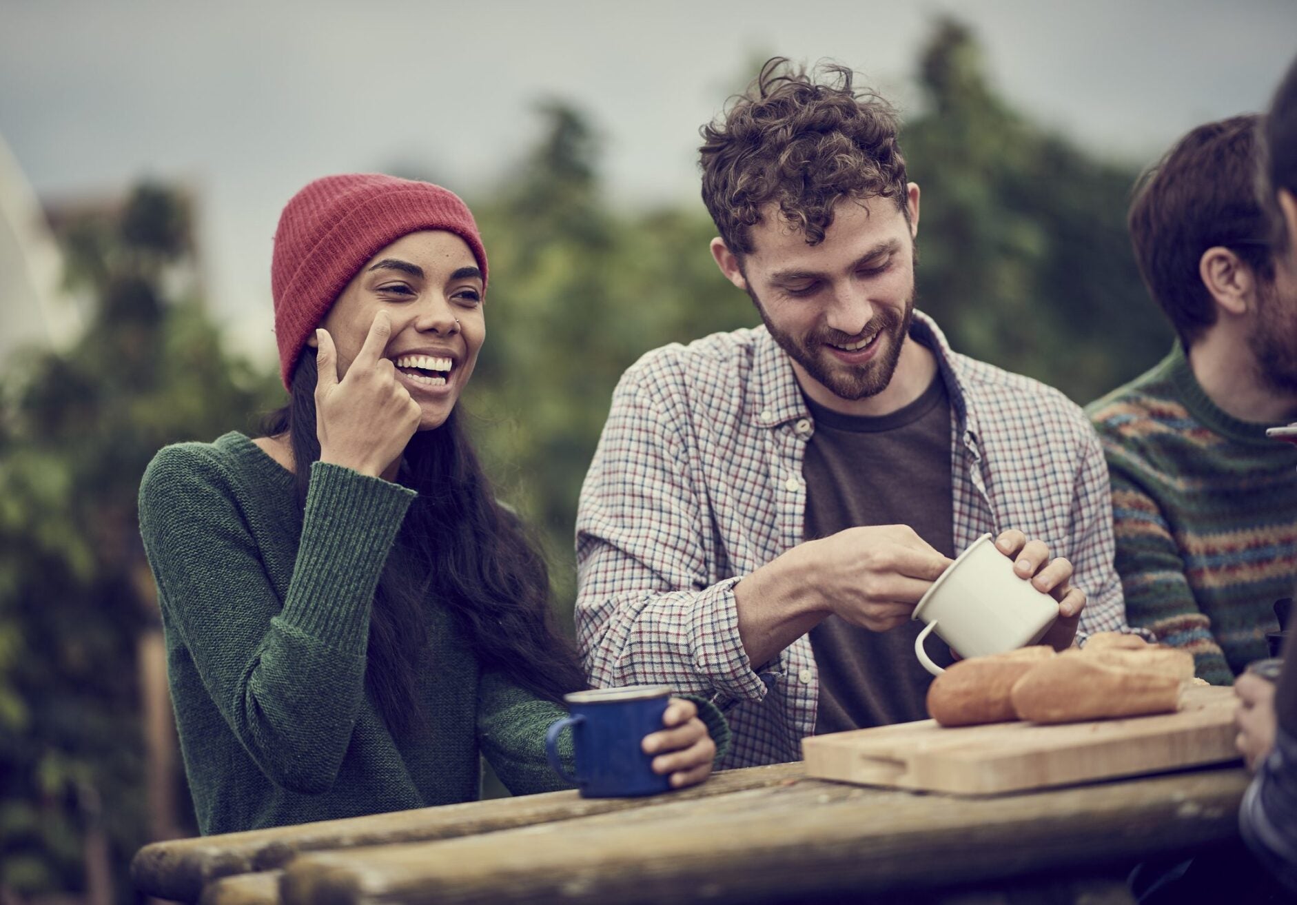 Farming friends taking a break from working in their community allotment, laughing and eating bread and soup together around picnic table