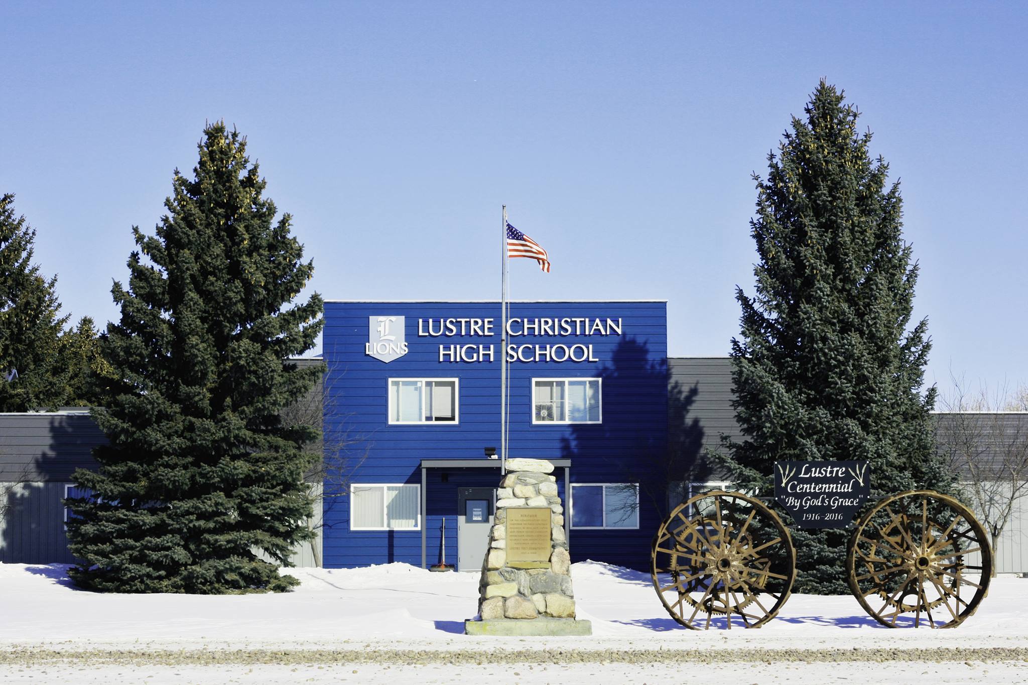 This is a picture of the front of Lustre Christian High School. The school is blue with white lettering with an American flag and wagon out front.